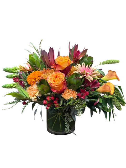 A vibrant autumn floral arrangement in a dark green cylindrical vase, featuring orange roses, yellow-orange calla lilies, pink and orange gerbera daisies, burgundy foliage, and green ferns, set against a plain white background.